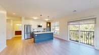 This contemporary single-floor residence showcases a thoughtfully designed open-concept layout with modern finishes throughout. The kitchen serves as a central highlight, featuring a large island with a blue-toned wood-grain finish and an integrated sink, complemented by pendant lighting above. Cabinetry combines crisp white upper cabinets with textured gray lower cabinets, set against a white subway tile backsplash. Stainless steel appliances, including a refrigerator, range, and over-the-range microwave, complete the space. Light hardwood-style flooring flows seamlessly from the kitchen into the open living area, which is bathed in natural light from large windows and a sliding glass door leading to a private balcony with outdoor views. The bedrooms feature plush carpeting, roller shades, and generous windows overlooking green surroundings. The bathroom is finished with large-format marble-style floor tiles, a glass-enclosed walk-in shower with a rainfall showerhead, and a wood-grain vanity. Throughout, the warm, neutral color palette and clean architectural lines create a bright, inviting ambiance that is both functional and refined.