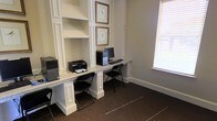 This single-floor business center offers a thoughtfully designed workspace ideal for professional tasks and everyday computing needs. The room features a built-in granite-topped workstation that spans the length of one wall, providing ample desk space for multiple users simultaneously. Two desktop computers and a printer are conveniently stationed along the counter, ensuring residents or guests have ready access to essential office equipment. Above the workstation, a set of open white shelving units offers organized storage space, while framed botanical artwork adds a refined, classic touch to the walls. The room is finished with elegant white wainscoting and panel molding that lend a timeless architectural character to the space. Dark carpeting grounds the room and contrasts neatly with the bright white millwork. A window fitted with blinds allows natural light to filter in while maintaining privacy, creating a comfortable and functional atmosphere. The overall ambiance is professional yet welcoming, balancing practicality with tasteful design details that make the space both efficient and visually appealing.