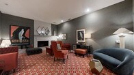Located at 737 Lockefield Ln in Indianapolis, Indiana, this thoughtfully designed lounge space offers a sophisticated and welcoming atmosphere well-suited for relaxation and social gatherings. The single-floor layout is anchored by a classic white brick fireplace, which serves as a focal point along one wall and lends a sense of warmth to the entire room. Deep charcoal-gray walls provide a rich, modern backdrop that contrasts beautifully with the bold terracotta and white geometric-patterned carpet, giving the space a striking visual energy. Seating is both stylish and comfortable, featuring plush rust-red wingback chairs paired with a matching ottoman, a tufted dark accent bench, and a blue curved accent chair that adds a complementary pop of color. Carefully selected décor elements — including diamond-shaped mirrors, framed bird artwork, and sculptural table lamps — contribute to a curated, design-forward ambiance. A dark wood console table along the far wall provides additional display and surface space. Recessed ceiling lighting ensures the room is well-illuminated while maintaining its intimate character. Overall, the space strikes a refined balance between bold contemporary design and inviting comfort.