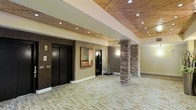 This single-floor lobby space combines rustic and modern design elements to create a welcoming and refined environment. The ceiling is a standout feature, clad in warm, natural wood planks accented by recessed pot lighting that casts a soft, even glow throughout the space. Structural stone columns add a rugged yet elegant architectural detail, grounding the space with a sense of permanence and craftsmanship. The walls are finished in neutral earth tones, complemented by subtle wall sconces that enhance the warm ambiance. Two sleek, black-finished elevator doors are prominently positioned, providing clean vertical access and contributing to the space's contemporary aesthetic. A framed piece of abstract artwork adds a curated touch to the wall, while a utility cart nearby hints at the space's functional role as a building lobby or common area. The flooring features a textured, neutral-toned carpet that ties the natural and modern elements together cohesively. The open layout offers a generous sense of space, allowing for easy movement and a comfortable, uncluttered flow. Overall, the lobby exudes a warm, upscale atmosphere with thoughtful design details throughout.