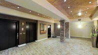 This single-floor lobby space combines rustic and modern design elements to create a welcoming and refined environment. The ceiling is a standout feature, clad in warm, natural wood planks accented by recessed pot lighting that casts a soft, even glow throughout the space. Structural stone columns add a rugged yet elegant architectural detail, grounding the space with a sense of permanence and craftsmanship. The walls are finished in neutral earth tones, complemented by subtle wall sconces that enhance the warm ambiance. Two sleek, black-finished elevator doors are prominently positioned, providing clean vertical access and contributing to the space's contemporary aesthetic. A framed piece of abstract artwork adds a curated touch to the wall, while a utility cart nearby hints at the space's functional role as a building lobby or common area. The flooring features a textured, neutral-toned carpet that ties the natural and modern elements together cohesively. The open layout offers a generous sense of space, allowing for easy movement and a comfortable, uncluttered flow. Overall, the lobby exudes a warm, upscale atmosphere with thoughtful design details throughout.