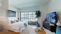 Located at 1050 Water St in the heart of Tampa's vibrant Water Street district, this thoughtfully designed studio offers a seamless open-plan layout that maximizes every inch of space. The main living area is anchored by a dramatic floor-to-ceiling sliding glass window wall that floods the interior with natural light and frames views of the outdoor pool and surrounding cityscape, creating a bright and airy atmosphere throughout.

Light-toned herringbone hardwood flooring flows continuously across the space, unifying the sleeping, living, and kitchen areas. A decorative lattice-style room divider provides a subtle visual separation between the sleeping zone and the living area, adding architectural interest without sacrificing the open feel. The kitchen is efficiently appointed with flat-front cabinetry in soft white and blue tones, stainless steel appliances including a built-in oven, microwave, and refrigerator, and a clean subway tile backsplash.

The bathroom features chevron-patterned tile flooring in warm earth tones, a glass-enclosed walk-in shower with subway tile surround, and a warm wood-framed vanity with an undermount sink. A built-in entry nook with a floating bench and wall sconce lighting serves as a charming and functional pet-friendly space. A walk-in closet completes this well-appointed urban retreat.