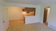 This single-floor space showcases an open-concept layout that connects the living area and kitchen, creating a cohesive and functional flow. The living area features neutral-toned wall-to-wall carpeting, contributing to a comfortable and understated atmosphere. Smooth white walls and a standard ceiling height give the space a clean, straightforward aesthetic.

The kitchen is defined by a half-wall breakfast bar that serves as a natural divider between the cooking and living zones while maintaining an open feel. Dark wood-toned upper cabinetry provides storage above the countertops, offering a contrast to the lighter walls and surfaces. The kitchen is equipped with white appliances, including a freestanding electric range and a refrigerator. An under-cabinet light above the range provides task lighting for the cooking area.

A vertical paneled accent wall element is visible near the kitchen, adding a subtle textural detail to the space. The overall design is practical and straightforward, with a neutral color palette throughout that offers a versatile backdrop. The single-floor layout ensures ease of movement and accessibility across the entire space.