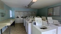 This communal laundry facility is designed to accommodate a high volume of use, offering a generous number of top-loading washing machines arranged in accessible rows alongside several stacked dryer units lining the back wall. The room's layout is thoughtfully organized to allow users to move freely between machines, making the laundry process efficient and straightforward. The walls are painted in a soft, muted blue-green tone, lending a calm and clean atmosphere to the space. Overhead, a ceiling fan provides air circulation, helping to maintain a comfortable environment during use. Lighting is provided by overhead fluorescent fixtures, ensuring the room is bright and well-lit throughout. The flooring is a neutral, easy-to-clean tile that complements the practical nature of the space. A folding or utility surface is visible along the perimeter, offering a convenient area for sorting and handling laundry. The overall ambiance is functional and well-maintained, reflecting a facility that prioritizes usability and convenience for its users. This single-floor laundry room serves as a practical, no-frills amenity built to meet the everyday needs of a shared residential or commercial setting.