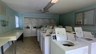 This communal laundry facility is designed to accommodate a high volume of use, offering a generous number of top-loading washing machines arranged in accessible rows alongside several stacked dryer units lining the back wall. The room's layout is thoughtfully organized to allow users to move freely between machines, making the laundry process efficient and straightforward. The walls are painted in a soft, muted blue-green tone, lending a calm and clean atmosphere to the space. Overhead, a ceiling fan provides air circulation, helping to maintain a comfortable environment during use. Lighting is provided by overhead fluorescent fixtures, ensuring the room is bright and well-lit throughout. The flooring is a neutral, easy-to-clean tile that complements the practical nature of the space. A folding or utility surface is visible along the perimeter, offering a convenient area for sorting and handling laundry. The overall ambiance is functional and well-maintained, reflecting a facility that prioritizes usability and convenience for its users. This single-floor laundry room serves as a practical, no-frills amenity built to meet the everyday needs of a shared residential or commercial setting.