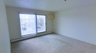 This single-floor space features a generously sized living area finished with light-colored wall-to-wall carpeting that adds warmth underfoot. The room is anchored by a large sliding glass door fitted with vertical blinds, allowing natural light to flood the interior while providing direct access to an outdoor area. Through the glass, views extend to a parking area framed by bare deciduous trees, offering a glimpse of the surrounding neighborhood. A baseboard heating unit runs along the exterior wall beneath the sliding door, providing an efficient heat source during colder months. The walls are painted in a clean, neutral tone, creating a blank canvas that complements a variety of furnishing styles. A wall-mounted unit, likely a thermostat or intercom panel, is positioned on the adjacent wall. The textured popcorn ceiling adds a subtle architectural detail consistent with the building's construction era. The overall ambiance of the space is clean, open, and functional, with an abundance of natural light making it feel airy and welcoming. The straightforward layout allows for flexible furniture arrangement to suit a range of needs.