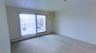 This single-floor space features a generously sized living area finished with light-colored wall-to-wall carpeting that adds warmth underfoot. The room is anchored by a large sliding glass door fitted with vertical blinds, allowing natural light to flood the interior while providing direct access to an outdoor area. Through the glass, views extend to a parking area framed by bare deciduous trees, offering a glimpse of the surrounding neighborhood. A baseboard heating unit runs along the exterior wall beneath the sliding door, providing an efficient heat source during colder months. The walls are painted in a clean, neutral tone, creating a blank canvas that complements a variety of furnishing styles. A wall-mounted unit, likely a thermostat or intercom panel, is positioned on the adjacent wall. The textured popcorn ceiling adds a subtle architectural detail consistent with the building's construction era. The overall ambiance of the space is clean, open, and functional, with an abundance of natural light making it feel airy and welcoming. The straightforward layout allows for flexible furniture arrangement to suit a range of needs.