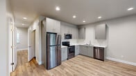 This two-story home offers a thoughtfully designed layout across two floors, providing comfortable living for everyday life. The main floor features an open kitchen and living area, where the kitchen stands out with its L-shaped configuration, gray shaker-style cabinetry extending to the ceiling, white countertops, and a full suite of stainless steel appliances including a refrigerator, electric range, over-the-range microwave, and dishwasher. Light wood-look flooring runs throughout the main level, complementing the neutral wall tones and recessed lighting overhead. A hallway with a carpeted staircase connects the floors, accented by clean white trim and warm, understated walls. The main floor also includes a bathroom and access to an attached garage with an automatic opener and an on-site water heater. Upstairs, two bedrooms offer comfortable retreats, including a generously sized primary bedroom featuring plush carpeting, multiple windows with views of the surrounding greenery, and a well-organized walk-in closet with built-in shelving and hanging rods. The second bedroom also features light hardwood-style flooring and natural light. Three bathrooms are distributed throughout the home to support daily routines with ease. A dedicated utility room adds practical functionality to the overall floor plan.