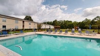 This outdoor amenity area centers around a generously sized swimming pool with clear turquoise water, enclosed by a black metal security fence that defines the space while maintaining open views of the surrounding lush tree line and manicured green lawn. The pool deck is finished in a smooth, light-toned concrete surface and is lined with multiple blue sling-style lounge chairs, providing ample space for sunbathing and relaxation.

Adjacent to the pool, a single-story clubhouse structure features large floor-to-ceiling glass windows and a glass-paneled entry door, offering direct visual access to the pool area from inside. The building's exterior is clad in light-colored vinyl siding, presenting a clean, low-maintenance aesthetic.

Beyond the pool, a wooden pergola provides a shaded gathering spot on the open lawn, while a colorful playground structure is visible near the perimeter fencing, making the area suitable for families. The combination of the spacious pool, dedicated sunning deck, shaded pergola, and recreational play area creates a versatile, well-rounded outdoor community amenity designed for leisure and social activity.