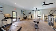 Located at 7925 Council Pl in Matthews, NC, this single-story fitness center offers a comprehensive workout environment thoughtfully organized into distinct exercise zones. The space is finished with durable patterned carpet tile flooring throughout and features recessed lighting complemented by natural light streaming through multiple large windows with blinds, providing views of the surrounding greenery.

The strength training area is visually anchored by a bold crimson accent wall, which contrasts with the otherwise light gray walls found throughout the facility. This zone houses a full dumbbell rack, a multi-function cable machine, adjustable benches, and additional strength equipment. A wall-mounted flat-screen television is positioned above the dumbbell station, and a water fountain is conveniently located nearby. Full-length mirrors reflect the space, enhancing the sense of openness.

A dedicated cardio area features a treadmill, elliptical trainer, and stationary bike, alongside a rack of medicine balls and foam rollers. A separate section accommodates additional selectorized strength machines, including seated and standing units. Motivational fitness artwork adorns the walls, contributing to an energizing atmosphere. Multiple entry and exit points, including glass-panel doors leading outdoors, ensure easy access throughout the facility.