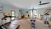 Located at 7925 Council Pl in Matthews, NC, this single-story fitness center offers a comprehensive workout environment thoughtfully organized into distinct exercise zones. The space is finished with durable patterned carpet tile flooring throughout and features recessed lighting complemented by natural light streaming through multiple large windows with blinds, providing views of the surrounding greenery.

The strength training area is visually anchored by a bold crimson accent wall, which contrasts with the otherwise light gray walls found throughout the facility. This zone houses a full dumbbell rack, a multi-function cable machine, adjustable benches, and additional strength equipment. A wall-mounted flat-screen television is positioned above the dumbbell station, and a water fountain is conveniently located nearby. Full-length mirrors reflect the space, enhancing the sense of openness.

A dedicated cardio area features a treadmill, elliptical trainer, and stationary bike, alongside a rack of medicine balls and foam rollers. A separate section accommodates additional selectorized strength machines, including seated and standing units. Motivational fitness artwork adorns the walls, contributing to an energizing atmosphere. Multiple entry and exit points, including glass-panel doors leading outdoors, ensure easy access throughout the facility.