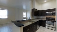 This single-floor space showcases a thoughtfully designed open-concept layout where the kitchen flows seamlessly into the living area. The kitchen is a standout feature, equipped with rich, dark espresso-toned cabinetry that extends both above and below the countertops, providing ample storage. The countertops are finished in speckled granite, lending a polished and durable surface throughout the workspace. A stainless steel sink is centrally positioned along the counter, accompanied by a modern gooseneck faucet. The kitchen also includes a peninsula with a matching dark countertop overhang, ideal for casual dining or additional prep space. A subtle tile backsplash adds texture behind the countertops. The adjoining living area benefits from a window that allows natural light to filter into the space, brightening the neutral-toned walls and light flooring. The ceilings are notably elevated, contributing to an open and airy atmosphere throughout. The light-toned flooring complements the dark kitchen elements, creating a balanced contrast. Overall, the space presents a clean, contemporary aesthetic with functional design details.