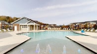 This single-level outdoor amenity space is designed to deliver a resort-like experience for its community. At its centerpiece is a generously sized swimming pool with a shallow wading entry and decorative water fountains that create an inviting, lively atmosphere. The pool deck is finished with aggregate concrete, providing both durability and a polished aesthetic, and is lined with lounge chairs ideal for sunbathing. A pool house structure, clad in board-and-batten siding, provides additional facilities and storage alongside the pool area. The surrounding residential buildings and rolling mountain ridgeline in the distance lend the space a picturesque, natural backdrop. Adjacent to the pool, a spacious covered patio extends from the main clubhouse building, which features warm brick exterior walls, white-trimmed windows, and white structural columns. Ceiling fans mounted overhead keep the covered area comfortable across seasons. This shaded lounge area is furnished with wicker seating — including a loveseat, armchairs, and ottomans with green cushions — arranged around a central coffee table, creating an inviting space for relaxation and socializing. The open concrete patio beyond offers additional al fresco dining with table-and-umbrella sets, completing a well-appointed outdoor gathering destination.