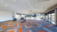 Located at 845 Yard St in Grandview Heights, Ohio, this single-floor fitness center is designed to deliver a premium workout experience in a bright, contemporary setting. The space is defined by expansive floor-to-ceiling windows that wrap around the perimeter, flooding the interior with abundant natural light and offering open views of the surrounding greenery — creating an energizing atmosphere for exercise. The clean white ceiling features recessed lighting and a ceiling fan, ensuring consistent illumination and comfortable airflow throughout. Underfoot, a bold herringbone-patterned carpet in shades of orange, gray, and blue adds a dynamic visual element that complements the modern aesthetic of the space. The fitness floor is generously proportioned, providing ample room between equipment stations for safe and comfortable use. A well-curated selection of strength and conditioning equipment is thoughtfully arranged throughout, including benches, cable machines, and resistance training stations. The open layout promotes ease of movement and accommodates multiple users simultaneously. Together, the abundant natural light, stylish finishes, and comprehensive equipment offerings make this fitness center a functional and visually engaging space suited for a wide range of workout routines.