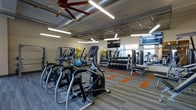 The Breakwater fitness center is a thoughtfully designed, single-floor workout facility that combines functionality with a modern aesthetic. The space features an open, airy layout anchored by an industrial-style ceiling with exposed ductwork, large-blade ceiling fans, and bright linear lighting that ensures the area is well-illuminated throughout.

The cardio zone is prominently positioned at the front of the space, housing a row of elliptical cross-trainers and other cardio equipment to accommodate multiple users simultaneously. Beyond the cardio area, a dedicated free weights section features a full rack of dumbbells alongside weight benches, offering a comprehensive strength training setup. Medicine balls and additional accessories are neatly organized along the walls for easy access.

Mounted flat-screen televisions are strategically placed to keep members entertained during workouts. The flooring features a neutral carpet tile with bold orange accent strips, adding a dynamic visual element while providing traction underfoot. Large windows allow natural light to flow into the space and provide views to an adjacent area, creating a bright, motivating atmosphere. The overall design balances practicality with a clean, professional environment suited for a wide range of fitness routines.