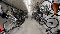 Located at 945-955 Reunion Avenue in Victoria, BC, this dedicated bicycle storage room is a single-level utility space offering a practical and organized solution for residents' cycling needs. The room features a concrete floor and grey-painted walls with an exposed, textured concrete ceiling overhead, giving the space a functional, utilitarian character typical of purpose-built storage areas.

Wall-mounted bicycle hooks and racks line both sides of the room, allowing bikes to be stored vertically and maximizing the available floor area. This dual-sided rack system creates a central aisle for easy navigation and access. A variety of bicycle types — including mountain bikes, road bikes, and commuter bikes — can be accommodated simultaneously, reflecting the room's high-capacity design.

A secured grey door with a small window at the far end provides controlled access to the space, reinforcing the safety and security of stored bicycles. Overhead piping and mechanical infrastructure run along the ceiling, consistent with a below-grade or ground-level utility environment. The layout is straightforward and efficient, designed entirely for function. This bicycle storage room represents a valuable amenity for cycling-oriented residents at this Victoria address.