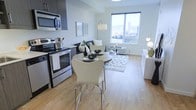 This contemporary single-floor residence showcases a thoughtfully designed open-concept layout that seamlessly integrates the kitchen, dining, and living areas into one cohesive, light-filled space. The kitchen is equipped with sleek stainless steel appliances, including a range, oven, and over-the-range microwave, all complemented by dark cabinetry that provides a striking contrast against the bright, neutral walls. A compact round dining table with modern white chairs sits just adjacent to the kitchen, making efficient use of the space while maintaining an airy feel. Light hardwood flooring flows throughout, adding warmth and continuity to the interior. The living area is anchored by a cozy sofa arrangement atop a patterned area rug, while a large picture window floods the room with abundant natural light and frames impressive panoramic views of a city skyline and surrounding mountains. The clean, minimalist aesthetic is carried throughout with contemporary furnishings, tasteful wall art, and carefully placed accent lighting. The overall ambiance is bright, modern, and inviting, making excellent use of the available footprint to create a functional yet stylish living environment.