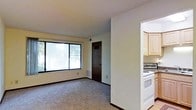 This single-floor residential space offers a straightforward and functional layout designed for comfortable everyday living. The living area is finished with wall-to-wall carpeting in a neutral tone, providing a cozy underfoot feel, and is illuminated by a large picture window that frames views of surrounding greenery and invites natural light deep into the room. A baseboard heating unit runs beneath the window, ensuring warmth throughout the space. The walls are painted in a clean, neutral off-white palette that keeps the interior feeling bright and open. A dark-toned interior door leads to an additional room or closet space, adding to the unit's overall functionality. The kitchen is accessible via an open arched passageway and is equipped with a white electric range and oven, light wood cabinetry providing ample storage, and a clean, practical layout suited for everyday meal preparation. The textured ceiling throughout adds a subtle architectural detail consistent with the unit's classic residential character. Overall, the space presents a clean, no-frills environment with all the essential features needed for day-to-day living.