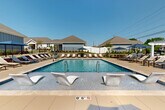 Located at 1702 Rocky River Road in Charlotte, NC, this outdoor amenity space centers around a generously sized, crystal-clear rectangular swimming pool designed for both relaxation and recreation. The pool features a shallow entry zone equipped with sleek, modern in-water lounge chairs, allowing residents to unwind while partially submerged — a standout resort-style detail. An accessibility lift is also present, ensuring the space is welcoming to all.

The expansive surrounding deck is finished in a warm, neutral tone and lined with a substantial number of slatted chaise lounges arranged to maximize sun exposure throughout the day. Shaded dining areas with market umbrellas and table seating provide a comfortable retreat from direct sunlight, ideal for outdoor gatherings. Raised landscaping beds filled with ornamental shrubs and flowering plants border portions of the deck, adding color and a sense of enclosure without sacrificing openness.

A charming hip-roof clubhouse structure with board-and-batten siding, French doors, and a pergola accent anchors one end of the pool area, serving as a functional and architectural focal point. The entire space is enclosed by a clean black iron fence, providing security while maintaining open sightlines to the surrounding neighborhood. Under Charlotte's abundant sunshine, this outdoor space offers a vibrant, resort-inspired atmosphere.