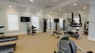 This fitness room offers a thoughtfully designed exercise environment finished with soft blue-gray walls, crisp white crown molding, and recessed lighting that creates a clean, polished aesthetic. The space is generously sized, providing ample room for a diverse selection of equipment including cardio machines, a cable weight system, a weight bench, and leg exercise equipment. Multiple wall-mounted flat-screen TVs are positioned throughout, allowing users to stay entertained during workouts. Two ceiling fans help maintain comfortable air circulation throughout the room. Natural light filters in through several white-trimmed windows, enhancing the open and airy atmosphere, while mirrored walls on one side visually expand the space and allow users to monitor their form. The neutral-toned carpet underfoot adds a layer of comfort and helps reduce noise. The overall design balances functionality with a refined, welcoming ambiance, making it a practical and pleasant environment for daily fitness routines.