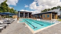 Located at 12801 Lucks Lane in Midlothian, Virginia, this two-story amenity center is a standout example of contemporary residential design. The building's exterior features a sleek combination of dark paneling and warm wood accents, anchored by dramatic oversized columns that frame floor-to-ceiling glass walls, seamlessly connecting the indoors to the outdoors.

At the heart of the space is a grand interior lounge boasting soaring double-height ceilings with warm wood detailing and a striking wall-mounted fireplace. Expansive glazed walls flood the room with natural light and offer unobstructed views of the resort-style pool deck. The lounge is furnished with stylish wicker and upholstered seating, creating an inviting, relaxed atmosphere.

The outdoor pool area is a central feature, with a large lap-style pool surrounded by a spacious stone-paved deck. Rows of chaise lounges, pergola-covered seating areas with navy blue umbrellas, and lush tree-lined surroundings contribute to a resort-like ambiance. An accessibility lift is also present at poolside. 

An outdoor terrace with café-style bistro seating bridges the interior and exterior zones, while a game room area equipped with a ping pong table, bar-height seating, and a wall-mounted display adds a recreational dimension to the facility.