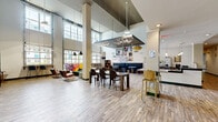 Located at 400 S Greene St in Greenville, NC, this impressive single-floor amenity space offers an extensive and thoughtfully designed environment for residents. The layout spans a generously sized footprint, encompassing multiple living areas, hallways, an exercise room, an office, and additional flexible spaces.

The main lounge area immediately commands attention with its soaring double-height ceilings, large industrial-style windows that flood the space with natural light, and wide-plank wood-look flooring. Pendant lighting and a stylish chandelier add a modern, curated aesthetic, while comfortable seating arrangements, a pool table, and eclectic wall art create an inviting social atmosphere. A community kitchen area features dark cabinetry, dark countertops, a full suite of appliances, and a bold geometric-patterned accent wall, making it ideal for gatherings.

Private office and meeting rooms are finished with carpeted floors, glass doors, built-in whiteboards, and round or oval conference tables, offering quiet, functional workspaces. A cozy booth-style nook with upholstered banquette seating provides an intimate spot for smaller meetings or study sessions.

The fitness center is well-appointed with high ceilings, sage-green walls, floor-to-ceiling mirrors, rubber flooring, and a comprehensive range of strength and cardio equipment. Hallways are accented with marble-tile flooring, vibrant artwork, and warm accent seating, tying the entire space together with a cohesive, modern residential feel.
