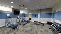 Located at 414 North Forest Park Boulevard in Knoxville, Tennessee, this fitness center offers a practical and well-appointed space designed to support a range of workout routines. The single-floor facility features a clean, modern interior with a neutral gray and blue color-blocked wall scheme that creates a focused, energizing atmosphere. Recessed lighting and a drop ceiling with integrated HVAC ensure a comfortable environment year-round. The space is equipped with cardio machines including a treadmill and an elliptical trainer, alongside strength and conditioning equipment such as a weight bench, medicine ball, and additional free weights. A wall-mounted flat-screen television is positioned for visibility from the workout area, adding an entertainment element during exercise sessions. The flooring consists of multi-toned carpet tiles in a patchwork pattern that adds texture and visual interest while providing a durable, easy-to-maintain surface. A mirrored interior window connects the fitness area to an adjacent room, enhancing the sense of openness. Natural light filters in through a window, contributing to the bright and inviting character of the space. Overall, the facility is thoughtfully organized to maximize functionality within a compact, accessible footprint.