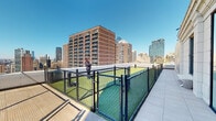 Located at 1401 Spruce St in the heart of Philadelphia, PA, this rooftop amenity level offers an exceptional outdoor experience with panoramic views of the city skyline, including recognizable landmarks such as the W Hotel tower and historic Philadelphia architecture. The space is thoughtfully designed to accommodate a variety of residents and their needs.

A generously sized, fenced dog run with artificial turf provides a safe and convenient area for pets to exercise, enclosed by chain-link fencing and easily accessible from the main terrace walkway. Adjacent to the dog run, a vibrant children's playground features colorful play structures and is fully enclosed with safety netting, offering a secure environment for young residents.

The open-air terrace features elegant large-format tile flooring and sleek glass panel railings that preserve unobstructed sightlines across the Philadelphia skyline. A louvered pergola structure adds architectural interest while providing partial shade to the outdoor seating areas, complemented by modern furnishings including stylish accent stools. The building's limestone-clad exterior walls frame large windows that look out onto the terrace, reinforcing the seamless connection between interior and exterior spaces. Bathed in natural light and open to clear blue skies, this rooftop deck serves as a dynamic, multi-use urban retreat above the streets of Center City Philadelphia.