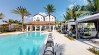 This outdoor amenity space showcases a striking Mediterranean-inspired design, centered around a generously sized swimming pool with crystal-clear blue water. The pool deck is finished in light-toned pavers, providing a clean and polished aesthetic that reflects the surrounding Florida sunshine. In-pool lounge chairs allow residents to relax directly in the water, while rows of cushioned chaise lounges line the deck for sunbathing.

To one side, a series of elegant private cabanas with sheer curtain panels offer shaded retreats, each equipped with seating for a more exclusive lounging experience. Tall, mature palm trees are strategically placed throughout the space, lending a tropical resort atmosphere while providing natural shade.

The backdrop is anchored by a multi-story white stucco clubhouse building featuring a red clay tile roof, arched colonnades, and a covered outdoor corridor — hallmarks of classic Mediterranean Revival architecture. The overall ambiance is sophisticated yet relaxed, combining upscale design elements with a vacation-inspired setting. The single-story pool pavilion connects seamlessly to the main building, creating a cohesive and inviting outdoor environment ideal for leisure and social gatherings.