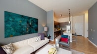 This thoughtfully designed single-floor residence offers a seamless blend of modern aesthetics and functional living. The open-concept layout connects the living area and kitchen, creating a bright and cohesive space ideal for both relaxing and entertaining. Soaring ceilings in the main living and kitchen areas add a striking sense of volume, while warm hardwood-style flooring flows throughout.

The living room is bathed in natural light from large windows fitted with horizontal blinds and sheer curtains, offering views of lush greenery outside. Bold gray accent walls and a private exterior door add character and convenience to the space.

The kitchen is a standout feature, anchored by a generous granite-topped island with bar seating. Dark espresso cabinetry provides ample storage, complemented by stainless steel appliances including a refrigerator, range, and microwave. Sleek pendant lighting illuminates the island and enhances the contemporary feel.

The bedroom is a serene retreat, carpeted for comfort and filled with natural light through a large window with sheer draping. The bathroom is well-appointed with a granite vanity, dark-framed mirror, wood-toned cabinetry, and a glass-enclosed tiled shower, completing this polished and cohesive residence.
