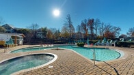 This two-floor outdoor amenity complex offers a well-appointed resort-style experience centered around a generously sized freeform swimming pool with a connected hot tub/spa. The pool area is finished with a paved brick-style surround deck, providing ample space for lounge chairs, teal umbrellas, and colorful seating arrangements. Multiple entry points with handrails make the pool accessible from various levels of the property.

A sweeping, manicured lawn area curves along the perimeter of the pool deck, lined with rows of teal and white lounge chairs and shade umbrellas, ideal for sunbathing and outdoor relaxation. The lawn is lush and well-maintained, offering a soft green contrast to the hardscape pool surround. Ornamental iron fencing encloses the entire complex, providing both security and a defined boundary while maintaining open sightlines to the surrounding tree-lined landscape.

A clubhouse-style building anchors one end of the property, featuring a covered porch and an additional covered cabana structure nearby. Mature deciduous trees frame the backdrop of the space, adding natural character throughout the seasons. The open-air layout is bathed in abundant natural sunlight, creating a bright, inviting atmosphere well-suited for outdoor leisure and community gatherings.