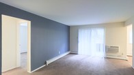 This single-floor space presents a clean and functional layout anchored by a spacious main living area. The room features a distinctive slate-blue accent wall that adds a touch of color and contrast against the surrounding neutral-toned walls and white ceiling. Soft, wall-to-wall carpeting in a warm beige tone covers the floor, contributing to a comfortable and quiet atmosphere underfoot.

A large sliding glass door dominates one wall, dressed with full-length vertical blinds that diffuse natural light evenly throughout the room. The door provides access to an outdoor area, and the light filtering through the blinds creates a bright, airy feel even when closed. The combination of the large glazed opening and light-colored ceiling enhances the sense of openness within the space.

Baseboard heating units run along the base of the walls, providing an efficient and unobtrusive heating solution. The overall design is straightforward and practical, with clean lines and minimal architectural detailing. The neutral palette and open floor plan offer flexibility for a variety of furniture arrangements and interior styles.