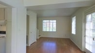 This two-story home offers a well-organized layout that balances practicality with comfort. The main living area immediately draws attention with its rich hardwood flooring that extends throughout, reflecting natural light and adding warmth to the space. Large windows along the exterior walls invite generous amounts of daylight, creating an open and airy atmosphere. The neutral, light-toned walls provide a clean and versatile backdrop, allowing the space to feel both welcoming and easy to personalize. A staircase connects the two floors, positioned conveniently within the living area to allow for smooth circulation throughout the home. Architectural details such as structural beams and clean white trim add subtle character to the interior. A closet door and floor-level air vent are also visible, pointing to thoughtful built-in storage and climate control features integrated into the design. The overall ambiance of the space is bright, understated, and move-in ready, offering a solid foundation for a variety of living arrangements. The two-floor configuration provides natural separation between common areas and private spaces, making this home well-suited for those seeking both open gathering areas and more secluded retreats.