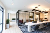 Located at 201 Infinity Way in Mountain View, California, this single-floor workspace delivers a polished, contemporary environment designed to support a variety of professional needs. The centerpiece of the space is an elongated, glossy white conference table surrounded by modern upholstered chairs, anchored beneath a striking tiered black pendant light fixture. Patterned area rugs and curated wall art add texture and visual interest throughout.

Floor-to-ceiling glass partitions with sleek black steel frames divide the space into distinct functional zones while maintaining an open, light-filled atmosphere. Natural light streams in through large windows, offering views of lush greenery outside. Private office areas enclosed by glass walls provide focused workspaces equipped with desks and task lighting, ensuring privacy without sacrificing the connected feel of the overall layout.

A unique highlight is the intimate booth seating area, featuring built-in cushioned benches, warm wood flooring, dark accent walls, and individual pendant lights — ideal for small meetings or informal collaboration. Clean white corridors with warm accent lighting connect the various zones, contributing to a smooth and professional flow throughout the space. The overall ambiance balances functionality with refined aesthetic sensibility, making it a well-appointed destination for modern professional work in the heart of Silicon Valley.