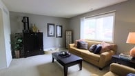 This single-floor space offers a welcoming and thoughtfully designed living area that balances comfort with style. The living room serves as the centerpiece of the home, finished with soft wall-to-wall carpeting in a warm neutral tone that complements the cool gray walls throughout. A large window allows generous natural light to fill the room, brightening the space and offering a view of the greenery outside.

The living room is furnished with a tan sofa accented by an assortment of decorative pillows in muted earth tones and navy blue, adding layers of texture and color. A dark wood coffee table serves as a practical and aesthetic focal point, styled with a decorative tray centerpiece. Alongside the seating area, a tall dark wood cabinet provides both storage and display space, topped with decorative vases and accents that contribute to the room's curated aesthetic.

Framed artwork on the walls and a decorative floor screen add visual interest and depth, giving the space a polished, intentional feel. The textured ceiling and clean lines of the room create a balanced, understated ambiance. Overall, this space presents a cozy yet refined environment ideal for everyday living and relaxed entertaining.