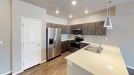 This single-floor residence offers a clean, modern design with a practical and open layout. The living area is anchored by rich, dark hardwood-style flooring that extends throughout the main living spaces, complemented by neutral gray walls and crisp white trim. The open-concept kitchen is well-appointed with dark espresso cabinetry, light quartz countertops, and a full suite of stainless steel appliances including a french door refrigerator with ice maker, electric range, and built-in microwave. A peninsula with pendant lighting provides additional counter space and casual dining options, opening naturally into the living area. The living room offers generous wall space and connects seamlessly to adjacent rooms through wide doorways. The bedroom features soft, neutral carpeting, a window fitted with blinds for light control, and ample space for furnishings. The bathroom is finished with a clean, white tub and shower combination with chrome fixtures. Throughout the residence, recessed and flush-mount lighting ensures consistent illumination, while the straightforward single-floor layout provides convenient, accessible living.