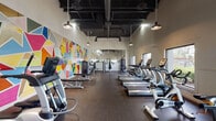 This fitness center is a thoughtfully designed, single-floor workout facility that combines functionality with striking visual character. The space is anchored by a large, eye-catching geometric mural spanning an entire wall, featuring a bold mosaic of colorful triangular and polygonal shapes that energizes the room and sets a dynamic tone throughout.

The layout is open and spacious, with soaring ceilings that enhance the sense of airiness. Exposed ductwork and dark-painted ceiling beams lend the space an industrial aesthetic, complemented by pendant-style hanging lights that provide warm, focused illumination across the floor. Durable rubber flooring runs throughout, offering both safety and practicality for high-activity use.

Cardio equipment is prominently featured, with a generous row of True-brand treadmills, ellipticals, and stationary cycling bikes arranged to allow comfortable spacing between users. Large windows along one wall invite natural light and provide views of outdoor greenery, creating a refreshing atmosphere during workouts.

The strength training area includes a selection of cable and resistance machines, adjustable benches, and a rack of dumbbells, catering to a wide range of training needs. A full-length wall mirror expands the visual depth of the space while allowing users to monitor their form. The overall ambiance is modern, vibrant, and motivating.