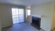 This single-floor space offers a comfortable and functional layout centered around a welcoming living area. The room's focal point is a classic fireplace framed by dark tile surround and a clean white mantle, adding warmth and character to the space. The hearth extends outward with matching tile, creating a defined and polished transition to the carpeted floor. Natural light floods the living area through a full-width sliding glass door dressed with vertical blinds, which opens to an outdoor balcony, offering views of the surrounding exterior. An additional window in the adjacent area further enhances the bright and airy atmosphere throughout. The walls are finished in a neutral, warm beige tone that complements the light-colored carpeting, contributing to an overall sense of openness. The textured ceiling adds a subtle architectural detail consistent with the space's practical and approachable design. The layout flows naturally between the living area and the adjoining space, making the floor plan feel cohesive and easy to navigate. Overall, this space provides a straightforward and livable environment with key comfort features, including a fireplace, outdoor access, and ample natural light.