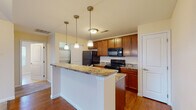 This single-floor residence at 1300 Community Way in Lancaster, Ohio offers a well-designed, open-concept layout that maximizes functionality and comfort throughout. The kitchen serves as a central highlight, featuring rich warm-toned cabinetry, granite-style countertops, and a peninsula island illuminated by a trio of pendant lights. Black appliances, including a range, built-in microwave, refrigerator, and double-basin sink, complete the kitchen's practical setup. Hardwood flooring runs throughout the kitchen and dining area, transitioning seamlessly into plush carpeting in the living and bedroom spaces. The living area benefits from vaulted ceilings that lend an open, airy feel, along with multiple windows dressed with blinds that invite natural light while maintaining privacy. A dedicated bedroom offers a quiet retreat with a window overlooking the exterior, while an additional carpeted room provides flexible use. The walk-in closet is outfitted with wire shelving and hanging rods, offering generous storage capacity. The bathroom includes a wood-toned vanity with countertop, tile flooring, a glass-enclosed shower, and a window that brings in natural light. Throughout, warm neutral wall tones and consistent finishes create a cohesive and inviting atmosphere.