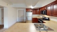 Located at 5055 William Flynn Highway in Gibsonia, PA, this single-floor unit offers a thoughtfully designed layout with a variety of functional and comfortable spaces. The living area is generously sized, featuring neutral wall-to-wall carpeting and a textured ceiling. Large windows dressed with vertical blinds flood the room with natural light and provide outdoor views of the surrounding greenery. A sliding glass door opens directly to a private balcony, extending the living space outdoors.

The kitchen is well-appointed with rich, dark wood cabinetry providing ample storage, light-toned countertops, and tile flooring. Stainless steel appliances include a refrigerator, gas range, and over-the-range microwave, offering a complete and modern cooking environment. A double basin sink with a view into the kitchen rounds out the practical design.

The bathroom features tile flooring and a warm wood vanity topped with a speckled granite countertop and an undermount sink. A large mirror paired with stylish industrial-style light fixtures enhances the space, while clean white walls maintain a bright, neutral ambiance throughout.

Additional carpeted rooms provide flexible space suitable for bedrooms or other uses, each with clean white walls and ceiling-mounted HVAC vents. The overall design is clean, neutral, and move-in ready.