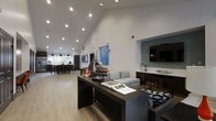 Located at 550 Harbor Cove Ln in Charleston, South Carolina, this single-floor community space offers a well-appointed, open-concept layout designed for both relaxation and social gatherings. The interior is anchored by striking vaulted ceilings that amplify the sense of openness and are fitted with recessed lighting, ensuring the space is bright and inviting throughout. Light hardwood-style flooring runs seamlessly across the entire area, contributing to a clean and cohesive aesthetic. The main lounge area features contemporary seating arrangements, a wall-mounted flat-screen television, and a built-in media console with dark cabinetry, creating a comfortable and functional focal point. Coastal-inspired decorative accents, including a model sailboat and nautical accessories, reflect the charming waterfront character of the Charleston locale. A dining area with bar-height seating and pendant lighting is visible toward the rear, offering an ideal setting for communal meals or casual gatherings. Colorful artwork displayed on the walls adds personality and a creative touch to the modern design. The overall ambiance is sophisticated yet welcoming, blending contemporary finishes with thoughtful coastal details that complement the spirit of the surrounding Harbor Cove community.