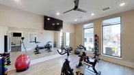 The yoga and fitness room is a versatile, single-floor space designed to accommodate a range of exercise routines. The room features light hardwood-style flooring that adds warmth while remaining practical for workouts. A full-length mirror spans one wall, visually expanding the space and providing a functional feature for form-checking during exercise. Recessed lighting in the ceiling, paired with a modern ceiling fan, ensures the room stays well-lit and comfortable during intense sessions. Large windows line one side of the room, allowing ample natural light to flood the interior while offering views of the surrounding outdoor environment. The space is equipped with multiple stationary spin bikes positioned throughout the room, along with a medicine ball rack stocked with a range of weighted balls for strength training. Stability balls and additional fitness accessories are visible, supporting diverse training needs. A large flat-screen television is wall-mounted near the mirrored wall, ideal for following guided workout sessions. The neutral, light-toned walls contribute to an open and energizing ambiance, making this a functional and inviting space for fitness enthusiasts.