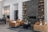 The Lobby Lounge in Caldwell, Idaho is a thoughtfully designed community space that blends contemporary style with warm, inviting materials across one generously proportioned floor. The grand entrance features soaring double-height ceilings adorned with warm wood paneling and dramatic pendant lighting, setting an upscale tone from the moment of arrival. Large-format tile flooring anchors the main entry and lounge areas, while light hardwood flooring flows through the interior gathering spaces. A standout stone-clad fireplace with flanking built-in shelving serves as the focal point of the main lounge, which is furnished with rich leather seating and a coffee table arrangement ideal for casual conversation. Floor-to-ceiling glazing and clerestory windows flood the interior with natural light and offer views of the outdoor pool and patio area. A coffee bar station, accented by a patterned tile backsplash and pendant lighting, provides a functional amenity hub. A dedicated lounge area features a tray ceiling with wood inlay, exposed beams, and a large-format media wall. The co-working and conference wing includes a formal conference room with textured wallcovering and a collaborative workspace with a long communal table, private offices enclosed by black-framed glass partitions, and abundant natural light throughout.