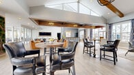 Located at 1038 Bayridge Dr in Lewis Center, Ohio, the Lake Club is a thoughtfully designed single-story community clubhouse spanning a generous footprint of open, well-appointed gathering space. The interior is anchored by soaring vaulted ceilings with exposed wood beam accents, lending a warm yet modern architectural character throughout. Light-toned hardwood flooring flows seamlessly across the main area, complementing the bright, airy atmosphere created by an abundance of large windows that invite natural light from multiple directions.

The main lounge area features a mix of seating zones, including leather bar-height tables and chairs, a relaxed lounge with cognac leather armchairs, and a cozy sitting nook furnished with patterned yellow accent chairs. A statement living area offers plush sofas, a round coffee table, and stylish drapery framing expansive outdoor views. A dedicated "Lake Club" branded bar and service counter with decorative tilework serves as a central focal point.

A fully equipped kitchen with slate-blue cabinetry, stone countertops, stainless steel appliances, and a mosaic tile backsplash provides functional support for events. A living wall installation adds a vibrant natural element. The space is finished with elegant chandelier lighting and a tray ceiling detail, creating a polished, welcoming ambiance throughout.