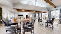 Located at 1038 Bayridge Dr in Lewis Center, Ohio, the Lake Club is a thoughtfully designed single-story community clubhouse spanning a generous footprint of open, well-appointed gathering space. The interior is anchored by soaring vaulted ceilings with exposed wood beam accents, lending a warm yet modern architectural character throughout. Light-toned hardwood flooring flows seamlessly across the main area, complementing the bright, airy atmosphere created by an abundance of large windows that invite natural light from multiple directions.

The main lounge area features a mix of seating zones, including leather bar-height tables and chairs, a relaxed lounge with cognac leather armchairs, and a cozy sitting nook furnished with patterned yellow accent chairs. A statement living area offers plush sofas, a round coffee table, and stylish drapery framing expansive outdoor views. A dedicated "Lake Club" branded bar and service counter with decorative tilework serves as a central focal point.

A fully equipped kitchen with slate-blue cabinetry, stone countertops, stainless steel appliances, and a mosaic tile backsplash provides functional support for events. A living wall installation adds a vibrant natural element. The space is finished with elegant chandelier lighting and a tray ceiling detail, creating a polished, welcoming ambiance throughout.