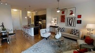 This single-floor residence showcases a thoughtfully designed open-concept layout that integrates the kitchen and living areas into one cohesive, contemporary space. The kitchen is outfitted with dark espresso cabinetry, stainless steel appliances, and a sleek breakfast bar with modern bar seating — ideal for casual dining or entertaining. Pendant lighting adds a refined touch above the counter, complementing the track lighting overhead. The living area exudes a modern aesthetic, anchored by warm hardwood flooring and a plush area rug that defines the seating space. A ceiling fan with a brushed metal finish provides both function and style. The room is furnished with a neutral-toned sofa accented by bold geometric throw pillows, paired with a contemporary white accent chair and a chrome and glass coffee table. A curated gallery wall featuring bold, graphic artwork adds personality and visual interest to the space. Decorative white vases and a console table with a table lamp complete the refined, layered look. The open flow between rooms creates a sense of spaciousness, while the cohesive color palette of neutrals, blacks, and bold red accents ties the entire space together with a polished, urban sensibility.