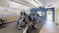 This fitness center offers a well-appointed and energizing environment designed to support a complete cardio workout experience. The space features soaring ceilings with exposed white steel roof trusses, lending an open, airy atmosphere that is further enhanced by abundant natural light streaming through multiple windows along the walls. The main workout area is outfitted with a comprehensive selection of Matrix-brand cardio equipment, including treadmills, elliptical trainers, upright stationary bikes, and recumbent bikes, all arranged to maximize usability and floor flow. The flooring transitions between dark commercial-grade carpet in the equipment zones and earthy tile in the circulation areas, adding visual definition to the space. A bold motivational wall graphic reading "Walk In Strong, Walk Out Stronger" greets visitors near the entrance, setting an encouraging tone. Large wall mirrors amplify the sense of space and allow users to monitor their form during workouts. A direct glass door provides convenient access to an outdoor pool area, with windows offering views of the surrounding grounds. Additional amenities include a water fountain station and accessible restroom facilities. A staff or check-in area is visible near the entrance, and the overall design balances functionality with a welcoming, community-oriented atmosphere.