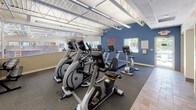 This fitness center offers a well-appointed and energizing environment designed to support a complete cardio workout experience. The space features soaring ceilings with exposed white steel roof trusses, lending an open, airy atmosphere that is further enhanced by abundant natural light streaming through multiple windows along the walls. The main workout area is outfitted with a comprehensive selection of Matrix-brand cardio equipment, including treadmills, elliptical trainers, upright stationary bikes, and recumbent bikes, all arranged to maximize usability and floor flow. The flooring transitions between dark commercial-grade carpet in the equipment zones and earthy tile in the circulation areas, adding visual definition to the space. A bold motivational wall graphic reading "Walk In Strong, Walk Out Stronger" greets visitors near the entrance, setting an encouraging tone. Large wall mirrors amplify the sense of space and allow users to monitor their form during workouts. A direct glass door provides convenient access to an outdoor pool area, with windows offering views of the surrounding grounds. Additional amenities include a water fountain station and accessible restroom facilities. A staff or check-in area is visible near the entrance, and the overall design balances functionality with a welcoming, community-oriented atmosphere.