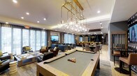 This stylish amenity lounge is designed to impress, offering a seamlessly connected layout that balances entertainment and relaxation across a single, open floor. At the heart of the space sits a full-size billiards table, positioned beneath a striking gold-framed chandelier adorned with cascading globe pendants that serve as a dramatic focal point. The open-concept design flows effortlessly from the game area into a comfortable lounge zone, furnished with plush blue sofas and warm leather armchairs arranged around a flat-screen television mounted on an accent wall. Floor-to-ceiling windows with sheer drapery flood the space with abundant natural light, enhancing the bright and welcoming atmosphere. Toward the rear of the lounge, a dedicated dining area features a round table surrounded by upholstered chairs, ideal for more intimate gatherings. Arched black architectural details and additional pendant lighting define this zone and add a refined contrast to the overall aesthetic. Recessed ceiling lighting throughout ensures the space is well-lit at all times. The combination of contemporary furnishings, luxurious lighting fixtures, and thoughtfully zoned areas makes this lounge a versatile and sophisticated destination for socializing and leisure.