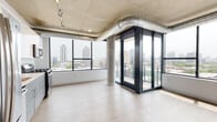 This contemporary urban residence showcases a striking industrial-modern aesthetic throughout. The main living area immediately impresses with its exposed concrete ceiling, polished visible ductwork, and structural concrete columns that lend an authentic loft-style character. Floor-to-ceiling windows wrap around the living space, flooding the interior with abundant natural light and framing expansive city skyline views. A distinctive glass-enclosed balcony vestibule with dark-framed sliding doors provides direct outdoor access while serving as a refined architectural focal point.

The kitchen is outfitted with sleek two-tone cabinetry — high-gloss white uppers paired with textured grey lowers — complemented by a white subway tile backsplash, quartz countertops, and a full suite of stainless steel appliances including a gas range, built-in microwave, and dishwasher.

The bedroom features light wood-style flooring, a dedicated walk-in closet, and direct access to a private ensuite bathroom, while exposed concrete above continues the industrial design language. Two well-appointed bathrooms serve the residence: one with a glass-enclosed walk-in shower with white subway tile and a modern vanity, and a second full bath with a tub-shower combination finished in matching tile. Light wood-style flooring flows consistently throughout, tying the clean, minimalist palette together.